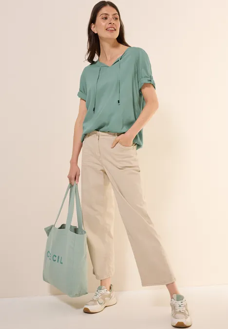 Frau in mintgrüner Bluse, beiger Hose und Turnschuhen, hält passende Tasche.